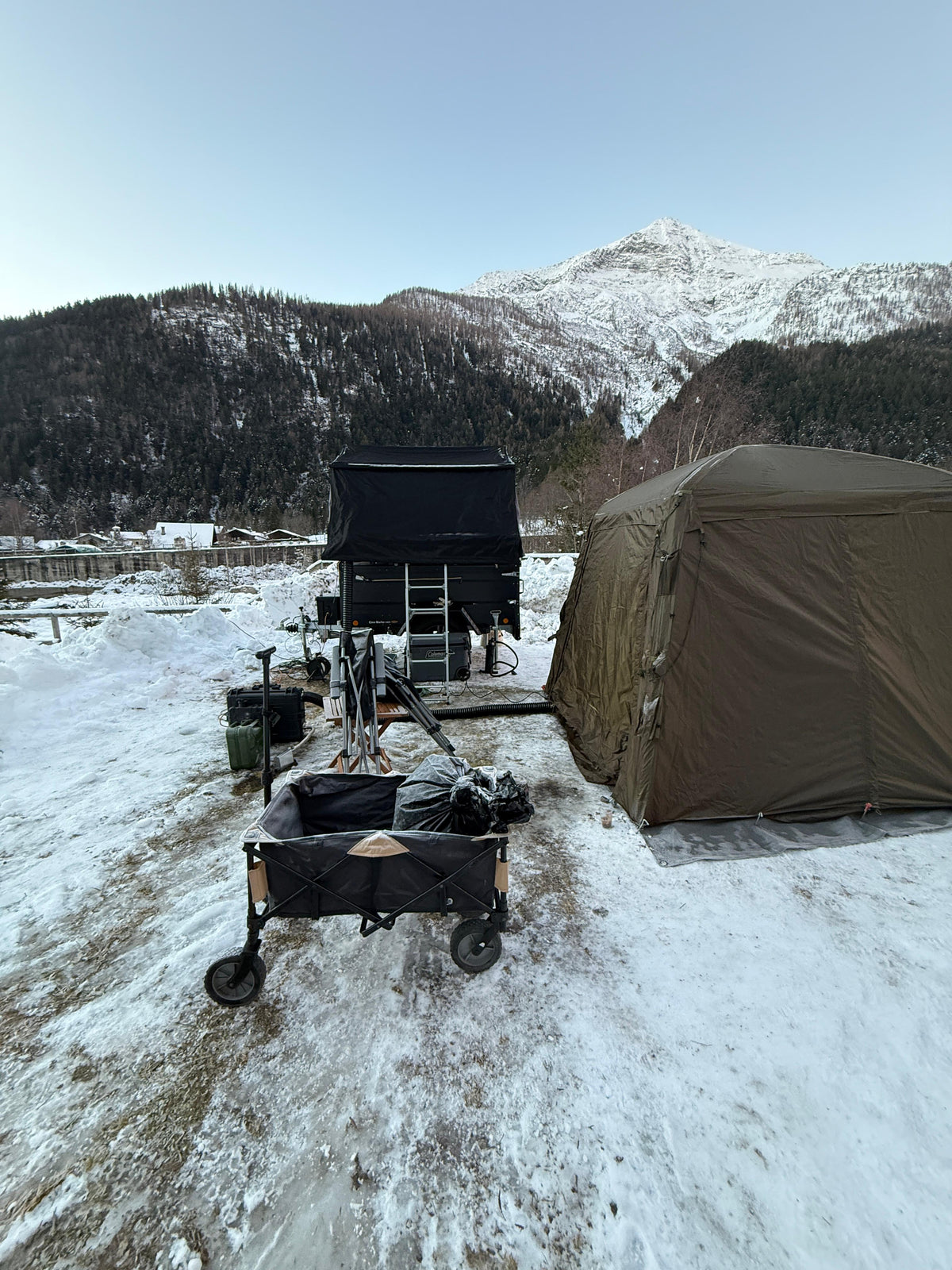 Campo invernale sulla neve con carrello tenda, tenda da tetto, tenda a terra e attrezzatura, montagne innevate sullo sfondo