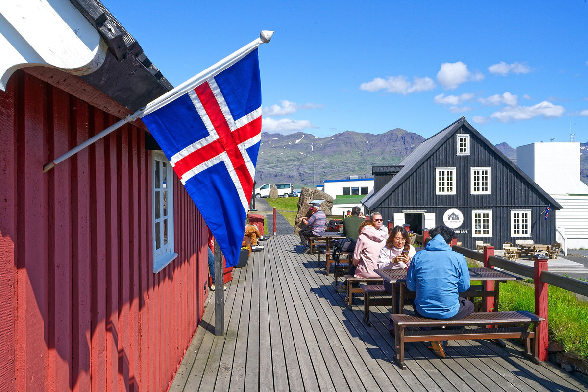Persone sedute ai tavoli all’aperto del caffè Langabúð a Djúpivogur con bandiera islandese