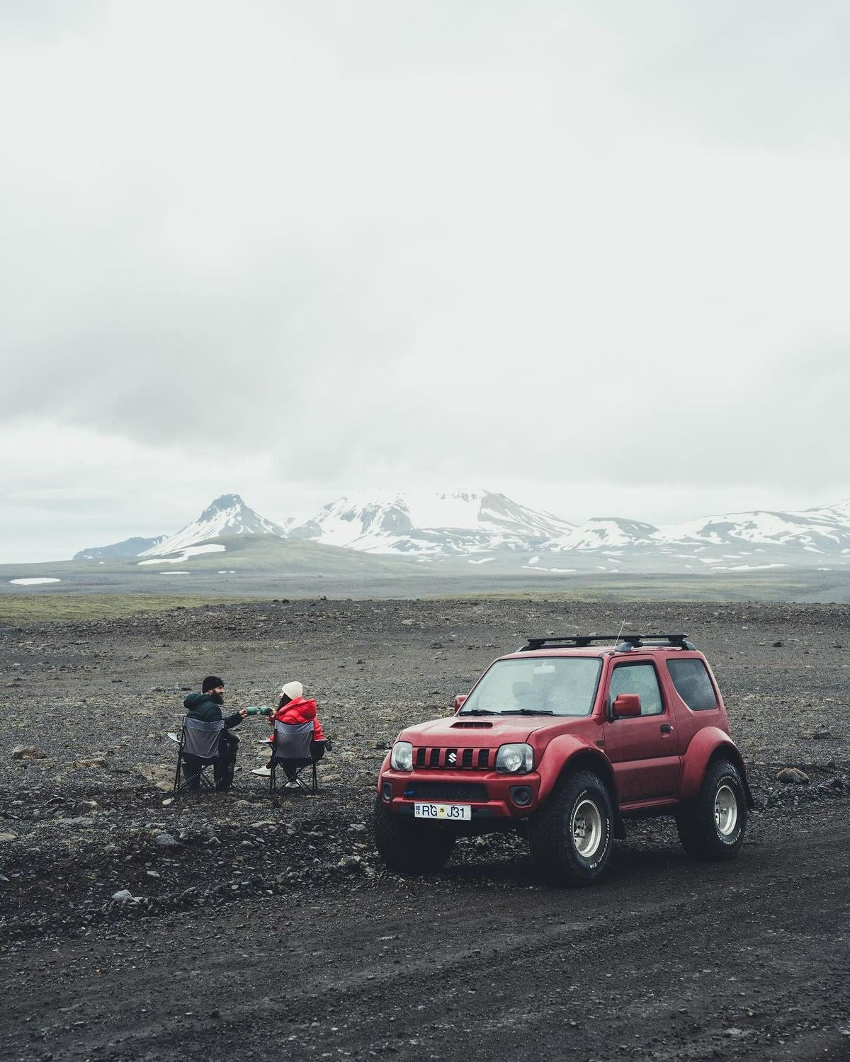 Suzuki Jimny rosso e due viaggiatori seduti su una piana nera delle Highlands